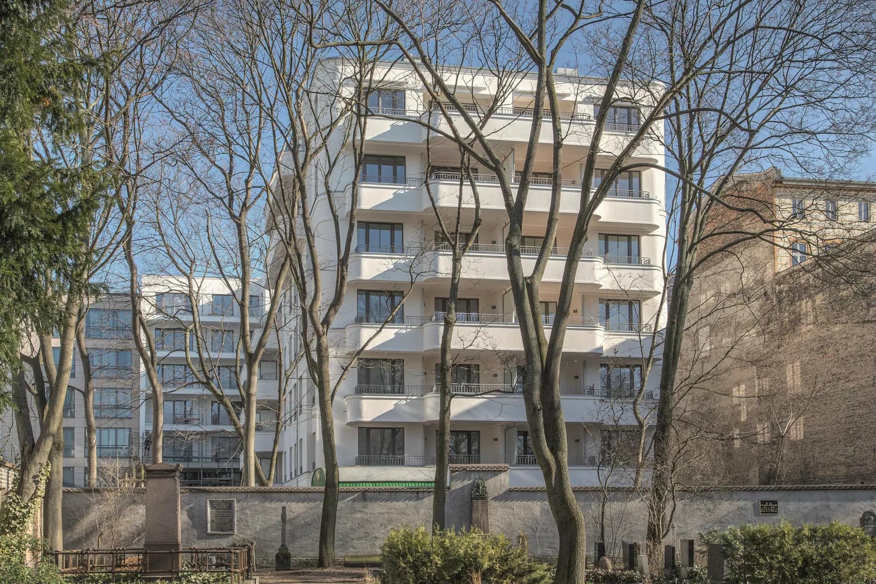 Rückseite des Seitenflügels mit geschwungenen weißen Balkonen, gesehen vom Dorotheenstädtischen Friedhof durch kahle Bäume.