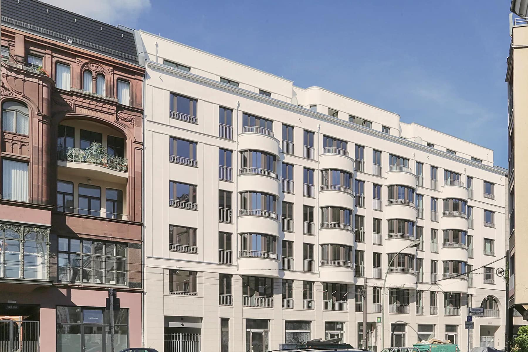 Straßenfront des weißen Neubaus an der Chausseestraße mit vier runden Erkerachsen, links der rote Sandstein-Altbau, im Vordergrund parkende Autos.